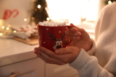 Photo of Woman holding cup of hot drink with marshmallow indoors, closeup