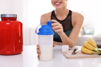 Young woman holding bottle of protein shake at table with ingredients in room, closeup