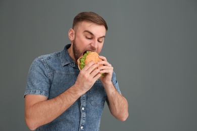 Photo of Young man eating tasty burger on grey background. Space for text