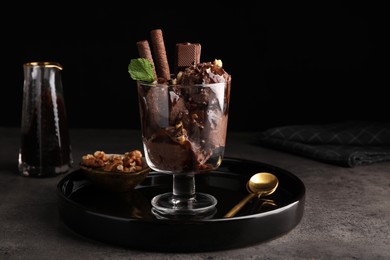 Photo of Tasty chocolate ice cream with sauce, nuts and wafer rolls in glass dessert bowl on dark grey table
