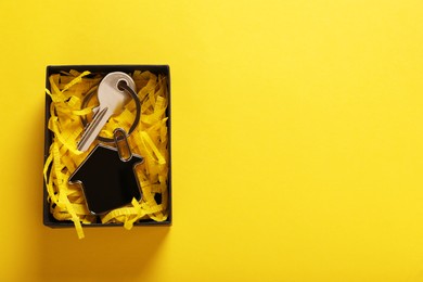 Photo of Key with trinket in shape of house and gift box on yellow background, top view with space for text. Housewarming party