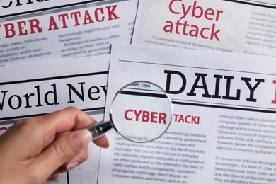 Photo of Woman using magnifying glass to read newspaper with headline CYBER ATTACK, top view