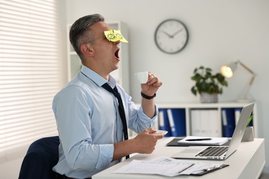 Photo of Man with fake eyes painted on sticky notes holding cup of drink and yawning at workplace in office