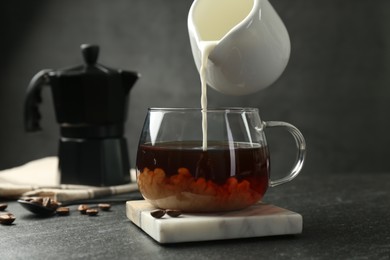 Photo of Pouring milk from pitcher into glass cup with coffee at dark textured table, closeup