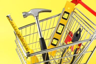 Photo of Small shopping cart with set of construction tools on yellow background, closeup