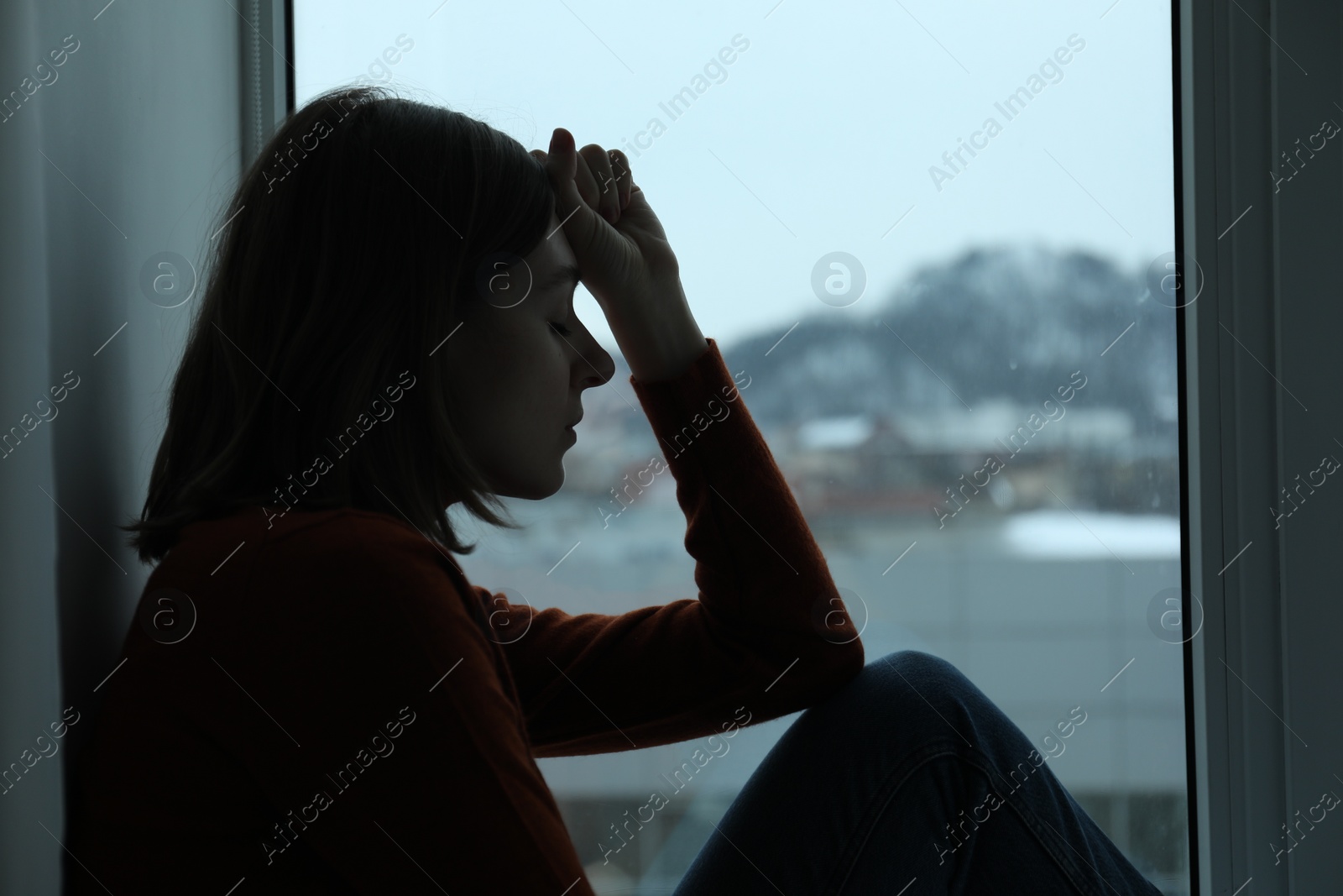 Photo of Sad young woman sitting on windowsill near window at home