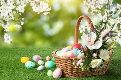 Image of Wicker basket with Easter eggs and beautiful flowers on green grass outdoors, space for text