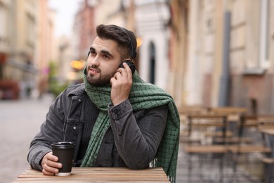 Photo of Handsome man in warm scarf with paper cup listening to music in outdoor cafe. Space for text