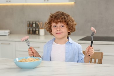 Photo of Cute little boy holding forks with sausages and bowl of pasta at table in kitchen