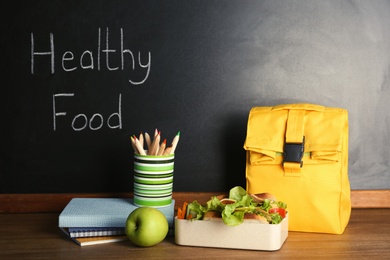 Photo of Lunch for schoolchild and stationery on table near blackboard with chalk written Healthy Food