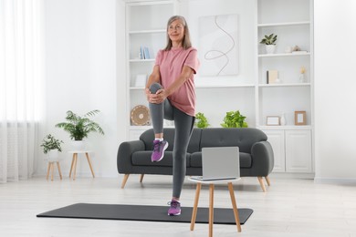 Photo of Senior woman in sportswear stretching near laptop at home
