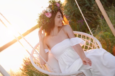 Photo of Young woman wearing wreath made of beautiful flowers on swing chair outdoors at sunset