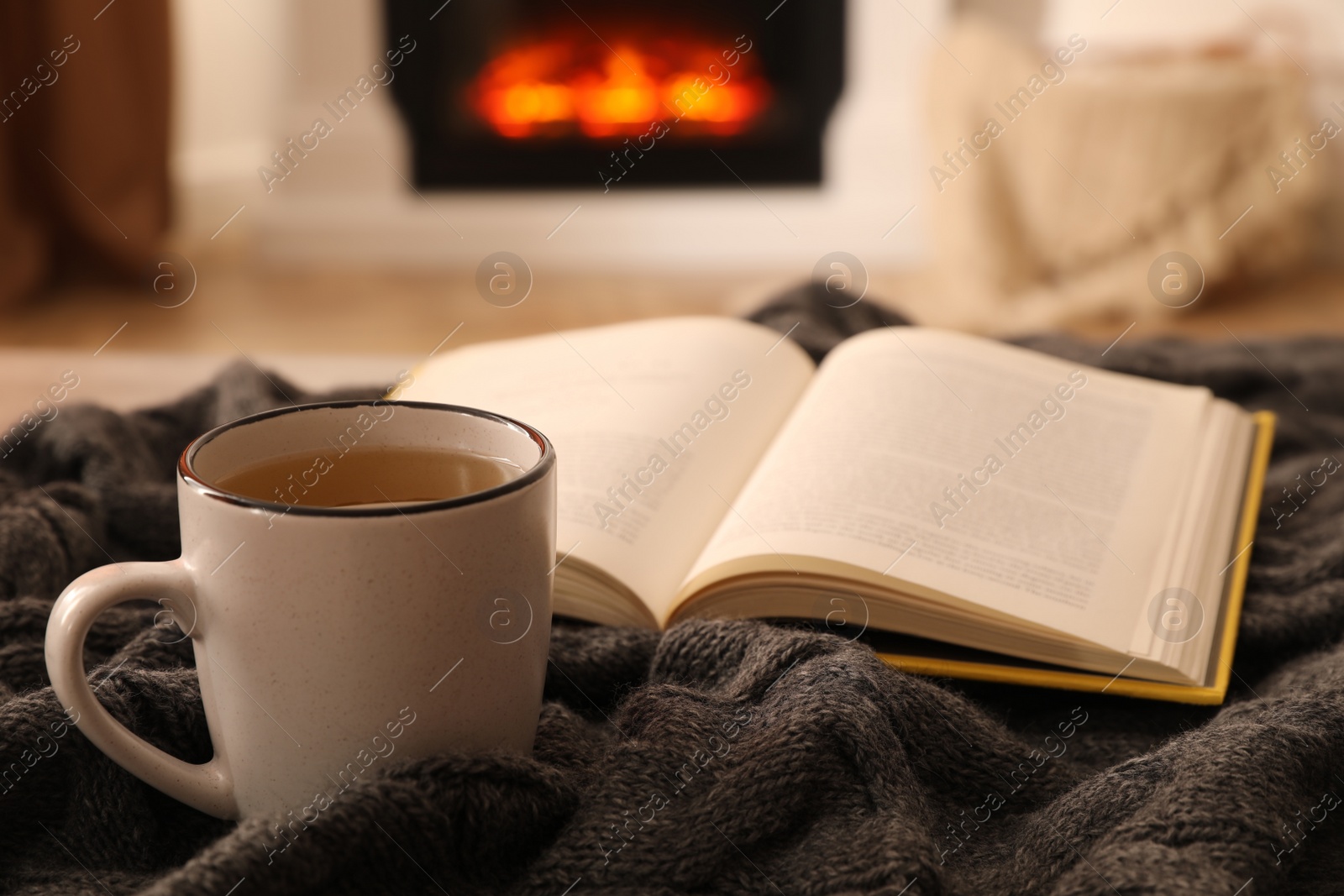 Photo of Cup of hot tea and book on knitted plaid near fireplace at home. Cozy atmosphere