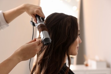 Hairdresser working with client using curling hair iron in salon, closeup