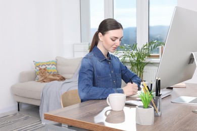Photo of Woman working at desk and cat lying on sofa in room. Home office