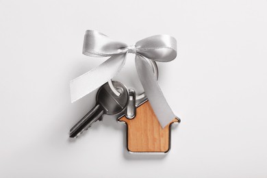 Key with trinket in shape of house and bow on light grey background, top view. Housewarming party