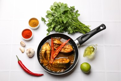 Tasty fish curry in frying pan and ingredients on white tiled table, flat lay. Indian cuisine