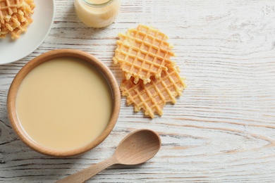 Bowl of condensed milk and waffles served on wooden table, top view with space for text. Dairy products