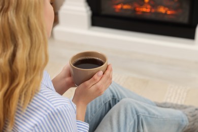 Photo of Young woman with cup of hot drink resting near fireplace at home, closeup