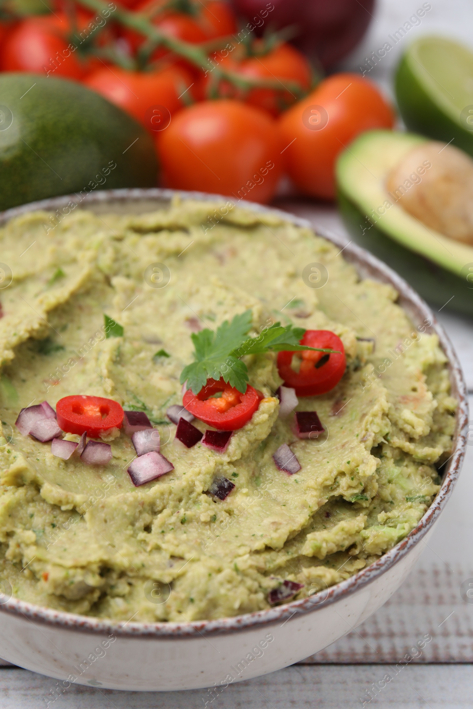 Photo of Bowl of delicious guacamole and ingredients on white wooden table, closeup