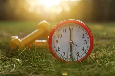 Photo of Alarm clock and dumbbells on green grass outdoors, closeup. Morning exercise