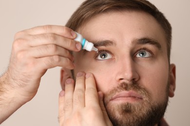Young man using eye drops on beige background, closeup