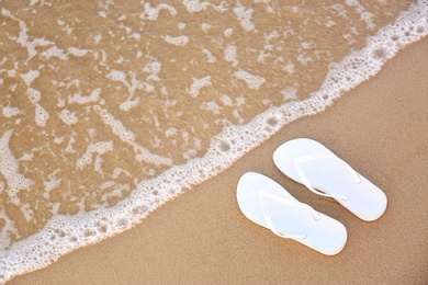 White flip flops on sand near sea, top view with space for text. Beach accessories
