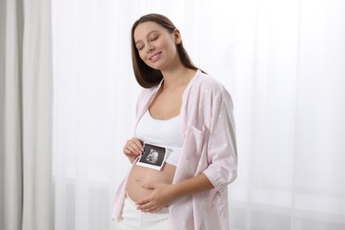 Beautiful pregnant woman with ultrasound picture of baby near window indoors