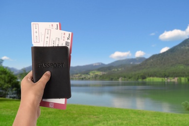 Image of Woman holding passport with tickets near lake and mountains, closeup. Travel agency service
