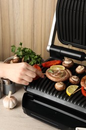 Photo of Woman cooking homemade sausages with mushrooms and bell pepper on electric grill at wooden table, closeup