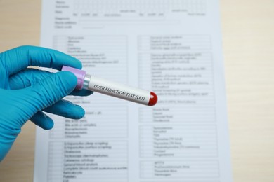 Photo of Laboratory worker holding tube with blood sample and label Liver Function Test over table, closeup