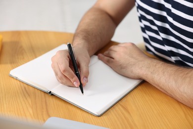 Photo of Young man writing in notebook at wooden table, closeup