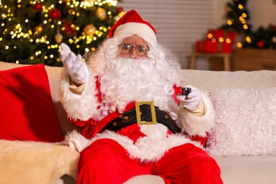 Photo of Merry Christmas. Emotional Santa Claus with remote control watching TV on sofa at home