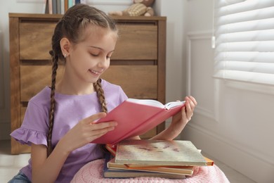 Photo of Cute little girl reading book near window at home