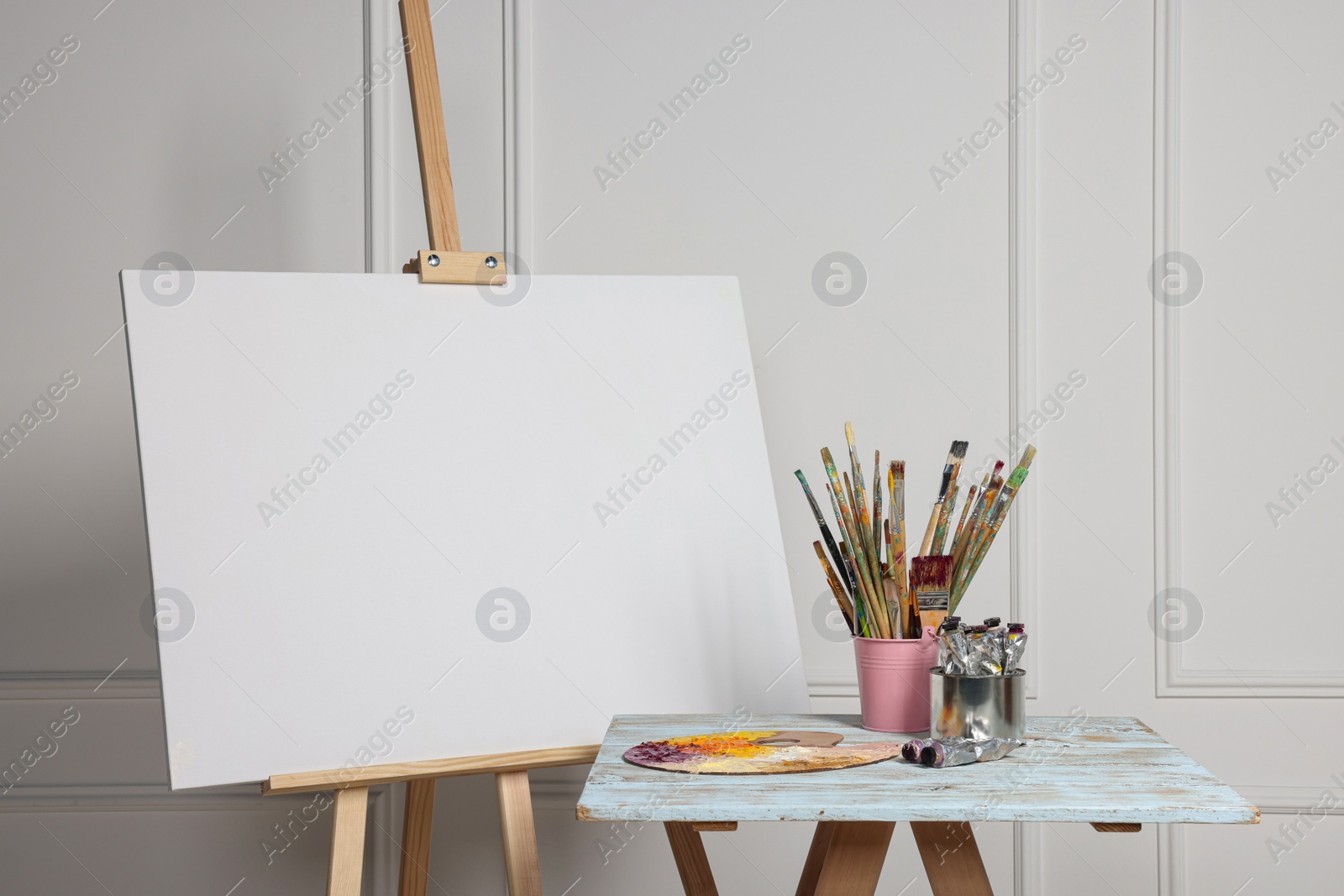 Photo of Easel with blank canvas and different art supplies on wooden table near white wall