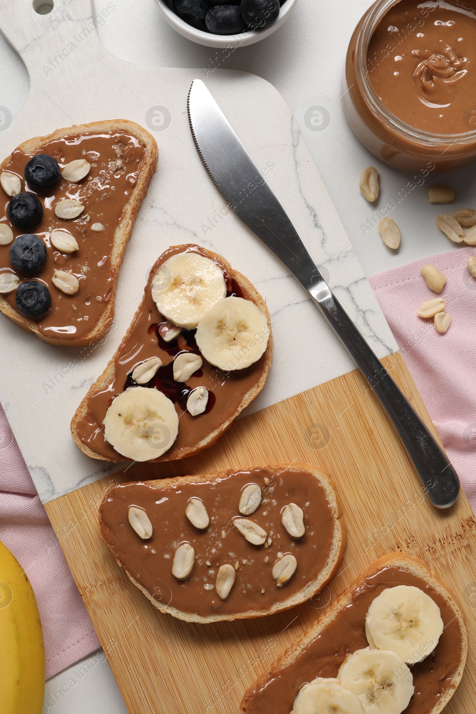 Photo of Toasts with tasty nut butter, banana slices, blueberries and peanuts on white table, flat lay