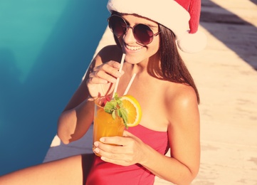 Photo of Young woman in Santa Claus hat with refreshing drink near swimming pool. Christmas vacation