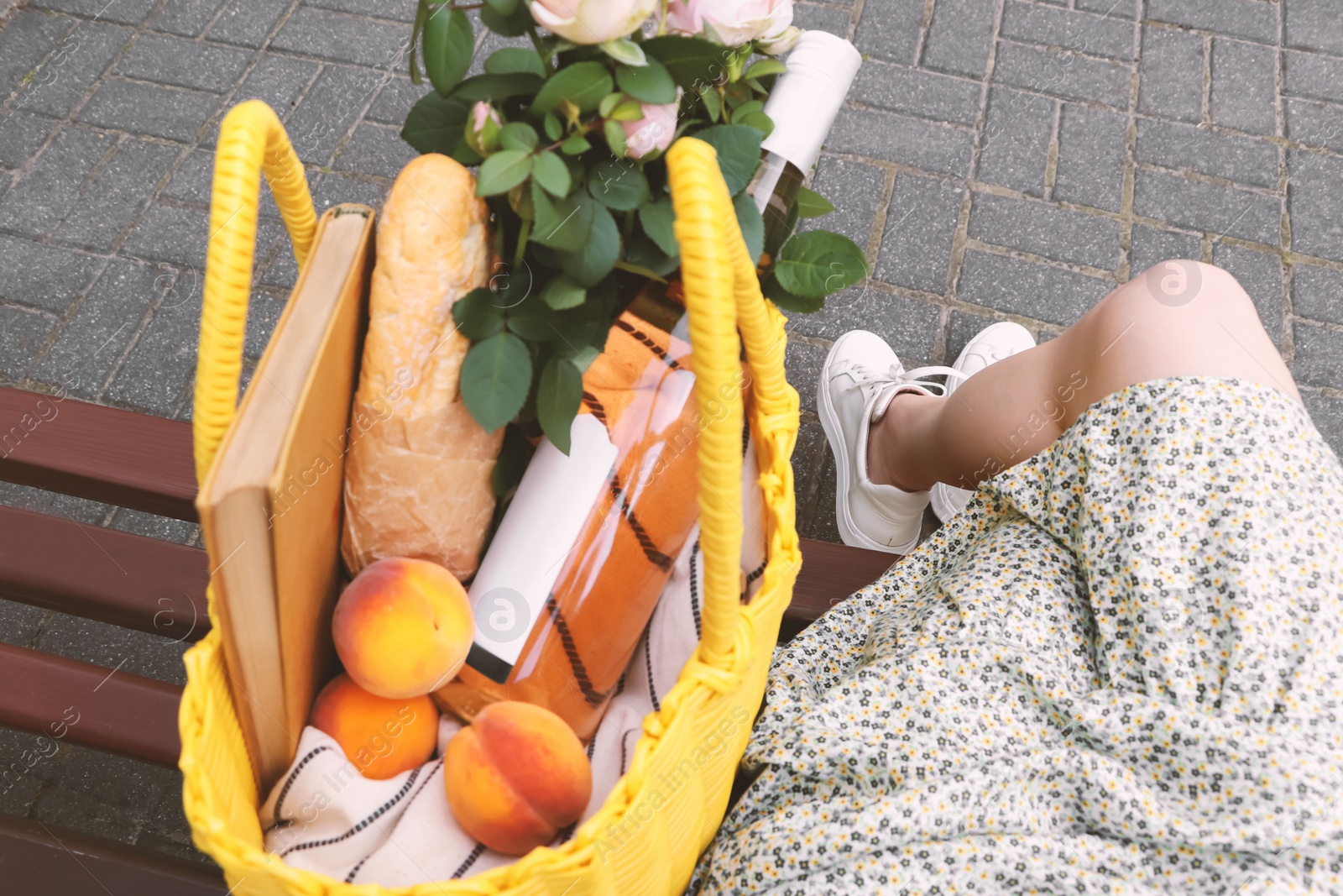 Photo of Woman sitting on bench near yellow wicker bag with roses, wine, peaches and baguette outdoors, closeup