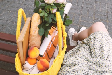 Photo of Woman sitting on bench near yellow wicker bag with roses, wine, peaches and baguette outdoors, closeup