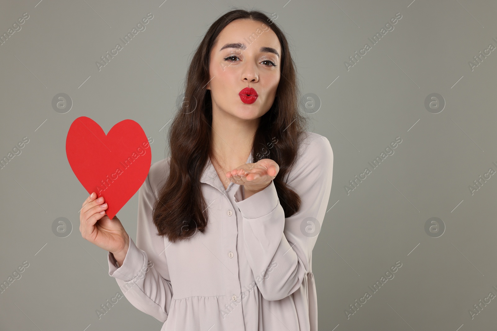 Photo of Beautiful young woman with paper heart blowing kiss on grey background, space for text