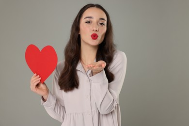 Beautiful young woman with paper heart blowing kiss on grey background, space for text