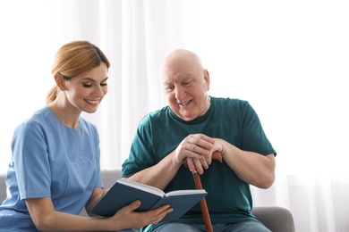 Photo of Nurse reading book to elderly man indoors. Assisting senior people