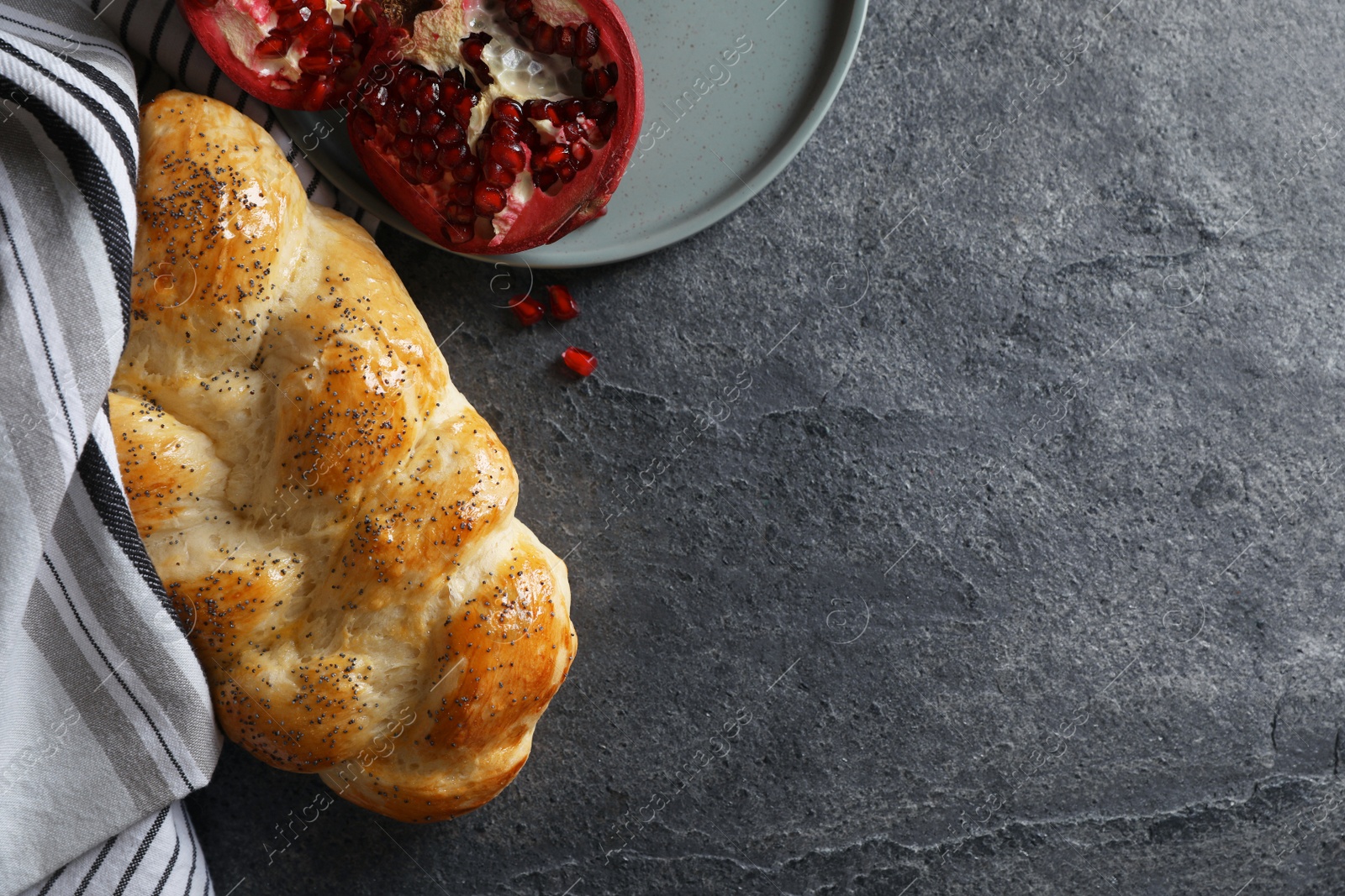 Photo of Homemade braided bread and pomegranate on grey table, flat lay with space for text. Cooking traditional Shabbat challah