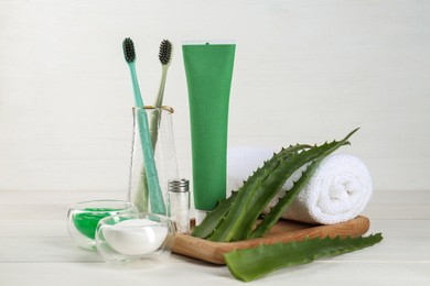 Tube of toothpaste, toothbrushes, fresh aloe vera leaves and care products on white wooden table