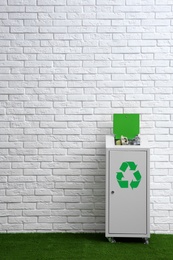 Overfilled trash bin with recycling symbol near brick wall indoors. Space for text