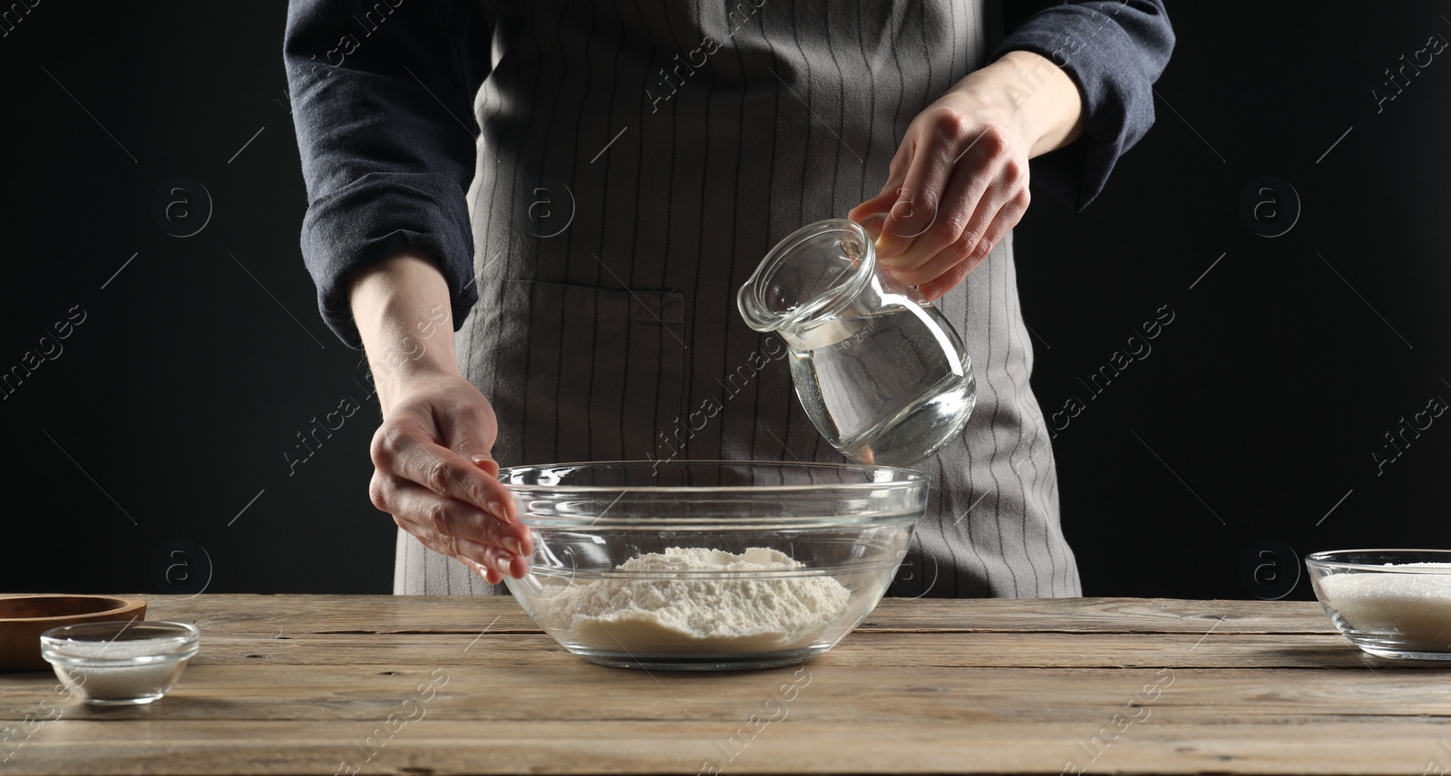 Photo of Making bread. Woman pouring water into bowl with flour at wooden table, closeup