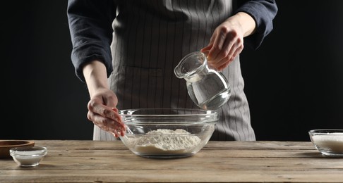 Photo of Making bread. Woman pouring water into bowl with flour at wooden table, closeup
