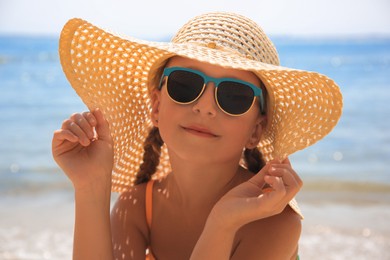 Happy little girl in stylish hat and sunglasses on beach near sea