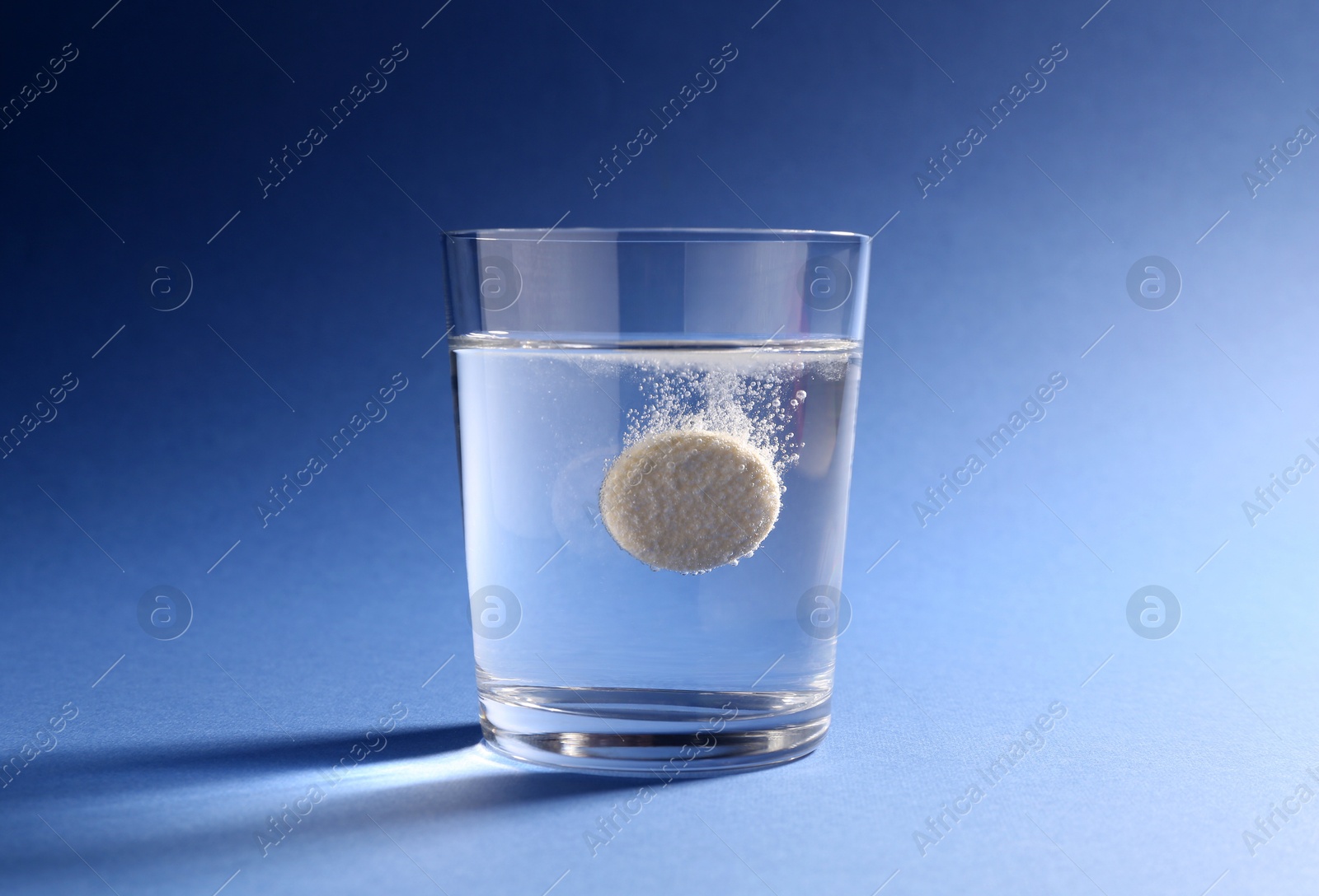 Photo of Effervescent pill dissolving in glass of water on blue background