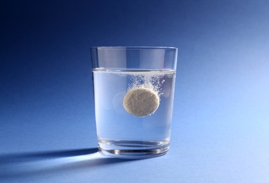 Photo of Effervescent pill dissolving in glass of water on blue background